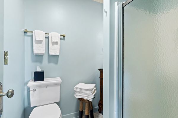 A small bathroom with a toilet, shower, neatly folded towels on a stool, and two towels on a rack above the toilet.