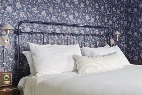 A bed with white pillows and sheets, a dark metal headboard, floral wallpaper, wall lamps, and a vintage radio on a bedside table.