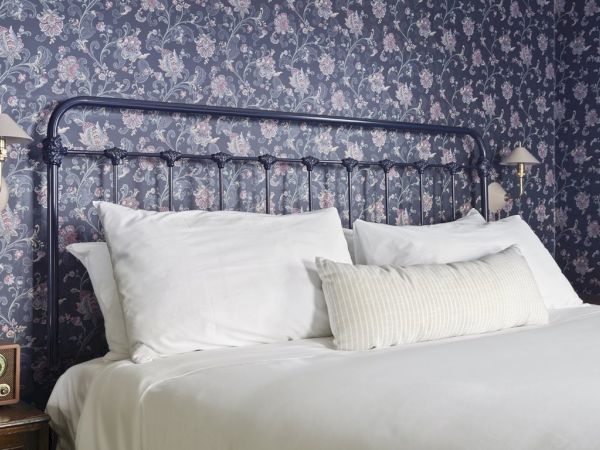 A bed with white pillows and sheets, a dark metal headboard, floral wallpaper, wall lamps, and a vintage radio on a bedside table.