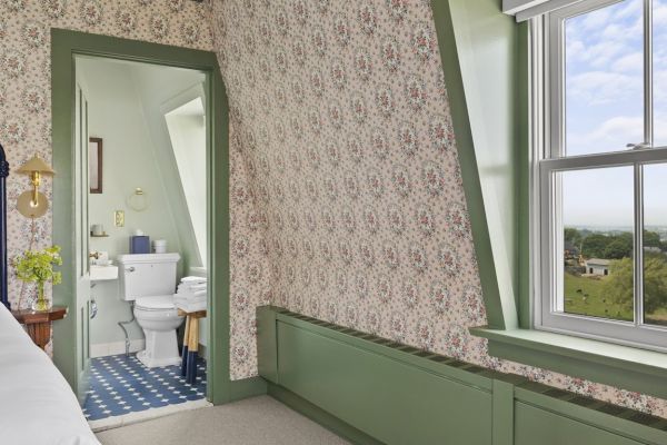 This image shows a bedroom with floral wallpaper, a bed, and a window. There's an adjacent bathroom with a blue and white tiled floor, sink, and toilet.