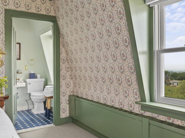 This image shows a bedroom with floral wallpaper, a bed, and a window. There's an adjacent bathroom with a blue and white tiled floor, sink, and toilet.