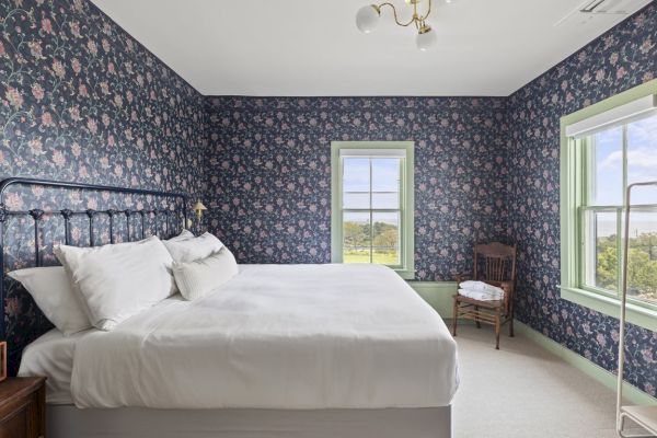 A bedroom with floral wallpaper, a large bed, green-trimmed windows, a chair, a bedside table with a radio, and a clothing rack.