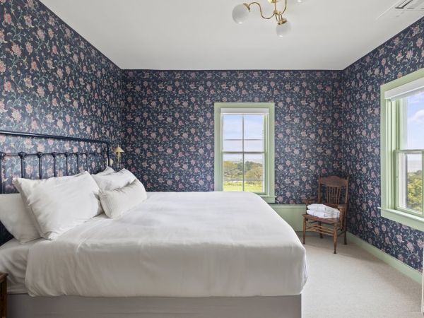 A bedroom with floral wallpaper, a large bed, green-trimmed windows, a chair, a bedside table with a radio, and a clothing rack.