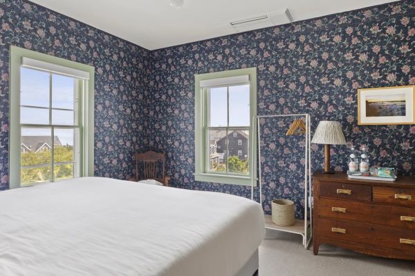 This image shows a bedroom with floral wallpaper, a bed, wooden dresser, lamp, framed picture, windows, a chair, and a freestanding coat rack.