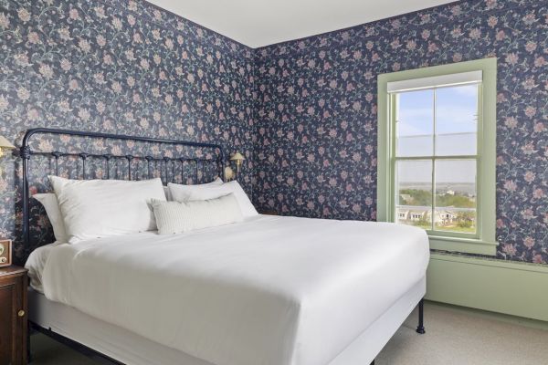 A cozy bedroom with floral wallpaper, a white bed, vintage bedside lamps, and a nightstand with a retro radio by a window with a view.