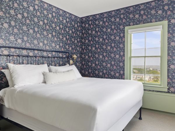 A cozy bedroom with floral wallpaper, a white bed, vintage bedside lamps, and a nightstand with a retro radio by a window with a view.