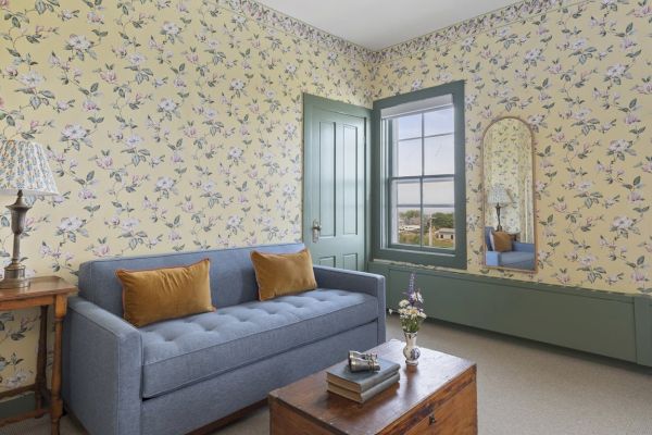 A cozy room with floral wallpaper, a blue couch with yellow cushions, a wooden coffee table, a vintage side table lamp, and a window with a view outside.