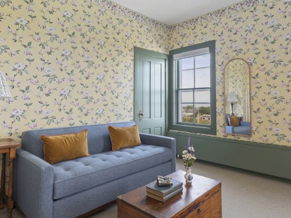 A cozy room with floral wallpaper, a blue couch with yellow cushions, a wooden coffee table, a vintage side table lamp, and a window with a view outside.