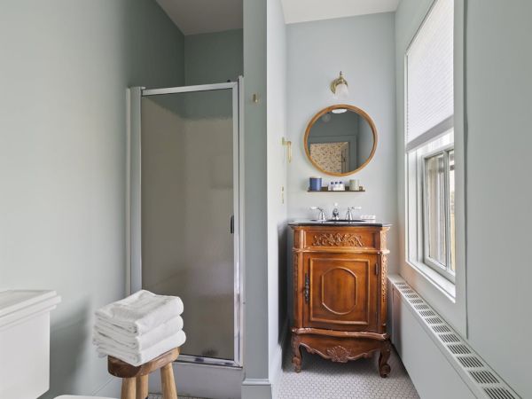 A bathroom with a glass shower, wooden vanity, round mirror, towels on a stool, and a window letting in natural light.