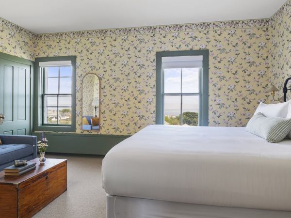 A cozy bedroom with floral wallpaper, a large bed, blue sofa, wooden chest, and two windows with a view. There is also a small coffee table.