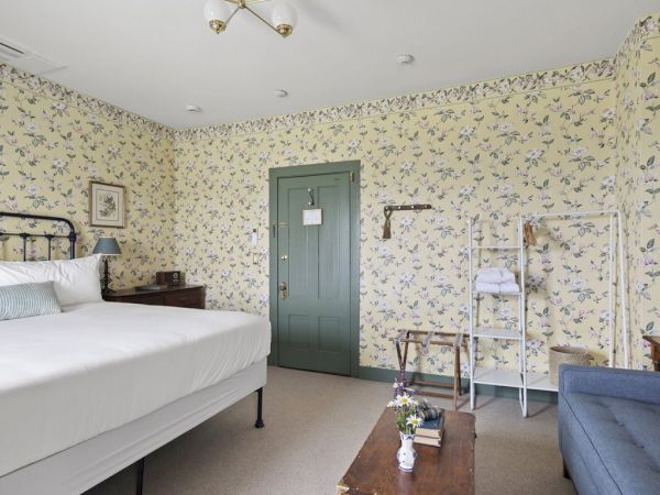 A cozy bedroom with a large bed, floral wallpaper, green door, sofa, coffee table, and clothes rack. The room has a vintage charm and ample lighting.