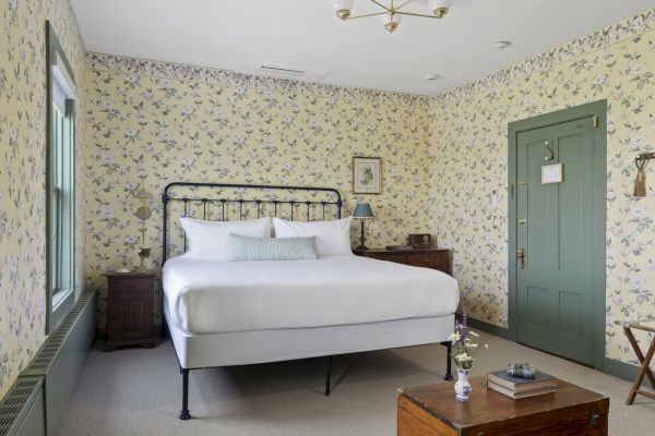 A cozy bedroom with floral wallpaper, a bed with white linens, wooden furniture, a green door, and a small table topped with books and flowers.