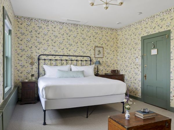 A cozy bedroom with floral wallpaper, a bed with white linens, wooden furniture, a green door, and a small table topped with books and flowers.