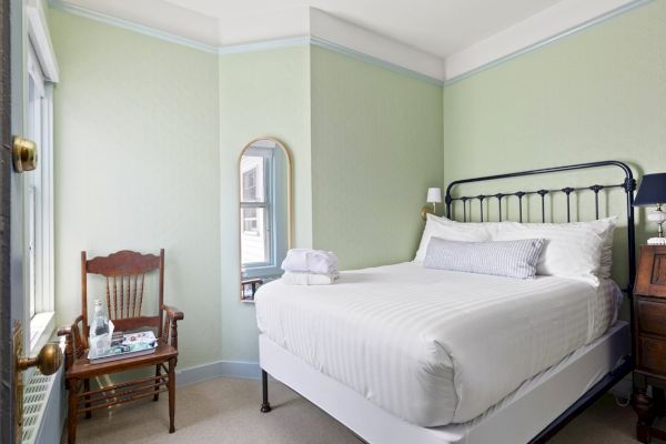 A small bedroom with a neatly made bed, a wooden chair with a tray, a mirror on the wall, a bedside table with a lamp, and a green color scheme.