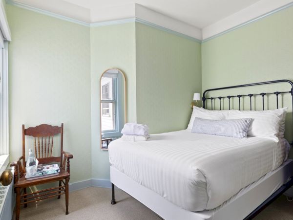 A small bedroom with a neatly made bed, a wooden chair with a tray, a mirror on the wall, a bedside table with a lamp, and a green color scheme.
