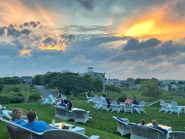 People are relaxing in chairs on a grassy hill during a picturesque sunset with scattered clouds in the sky.