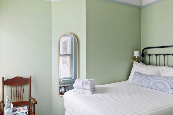 A cozy bedroom with a green accent wall, a metal-frame bed with white linens, a wooden chair by a window, and a small mirror on the wall.