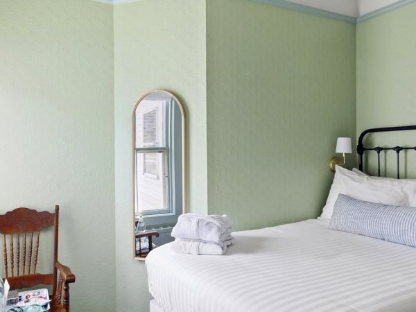 A cozy bedroom with a green accent wall, a metal-frame bed with white linens, a wooden chair by a window, and a small mirror on the wall.