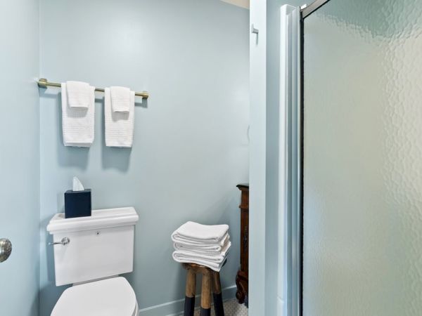 A bathroom with a toilet, towel rack holding two white towels, a tissue box, a stack of towels on a stand, and a frosted glass shower door.