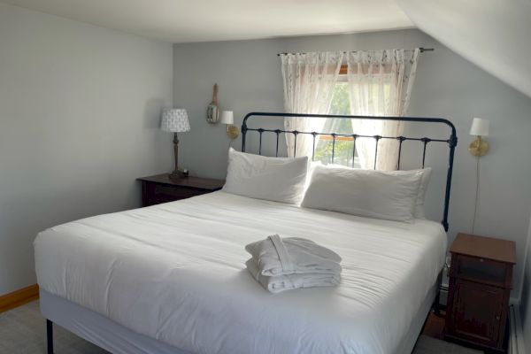 A bedroom with a neatly made bed, folded towels on it, two side tables, lamps, and a window with white curtains.