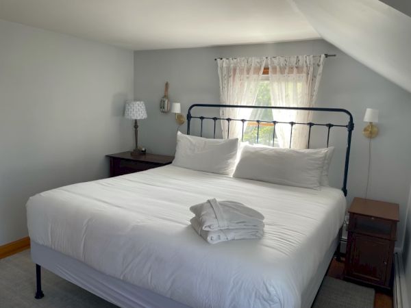 A bedroom with a neatly made bed, folded towels on it, two side tables, lamps, and a window with white curtains.