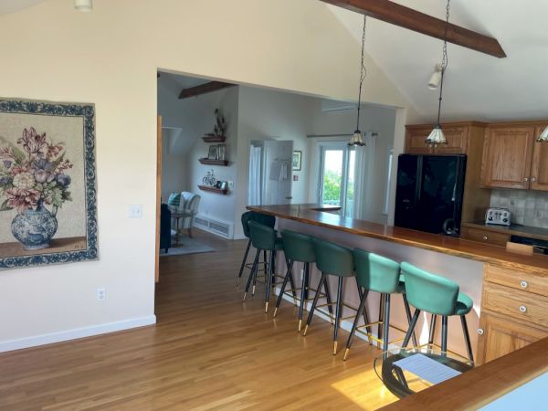 This image shows a kitchen area with a bar counter, green bar stools, wooden flooring, hanging lights, and a framed floral artwork on the wall.