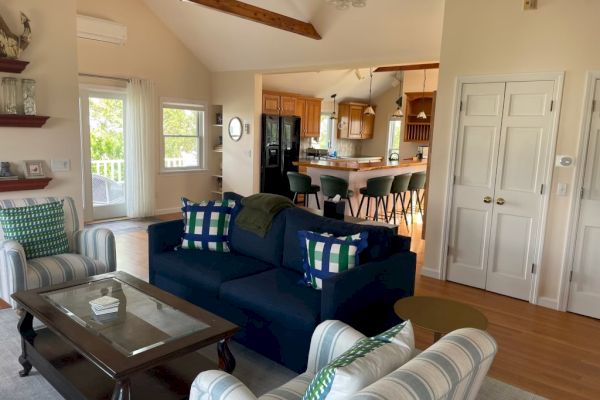 A cozy living room features a dark blue sofa, armchairs, and a coffee table. It opens to a bright kitchen with a dining area and bar stools, ending the sentence.