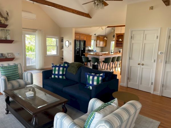 A cozy living room features a dark blue sofa, armchairs, and a coffee table. It opens to a bright kitchen with a dining area and bar stools, ending the sentence.