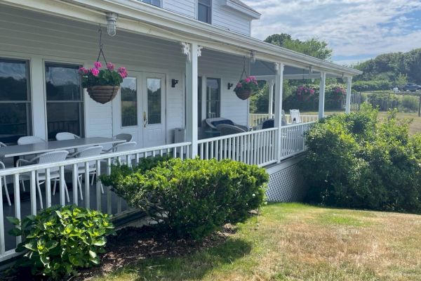 A porch with hanging flower baskets and an outdoor seating area attached to a white house, surrounded by shrubs and an open grassy area.