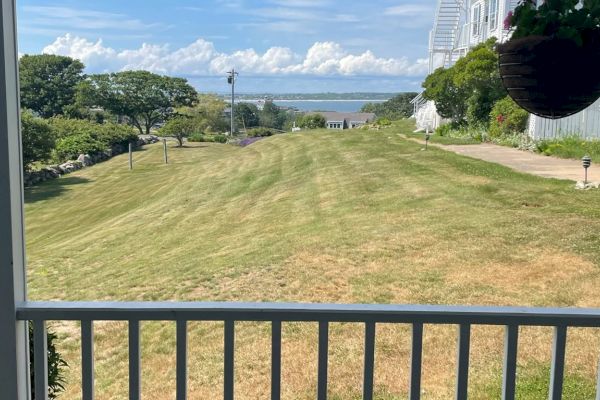 A clear sky over a grassy lawn leading to a distant bay, viewed from a porch with a white railing and a hanging plant.