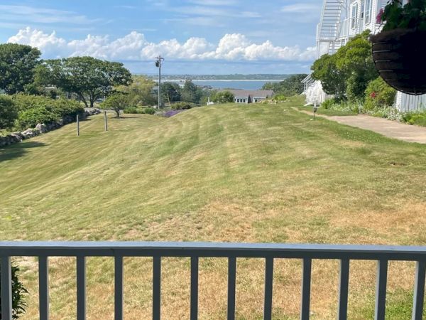 A clear sky over a grassy lawn leading to a distant bay, viewed from a porch with a white railing and a hanging plant.
