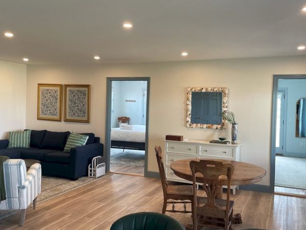 A cozy, furnished living room with a blue sofa, armchairs, a dining table, and wall art, leading to other rooms through doorways.