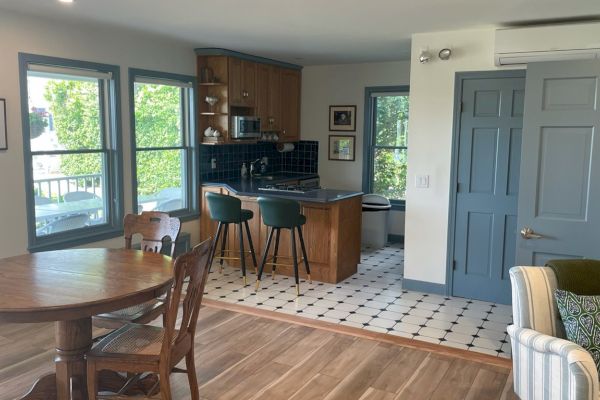 A cozy kitchen and dining area featuring wooden furniture, green bar stools, a small island, and large windows bringing in natural light.