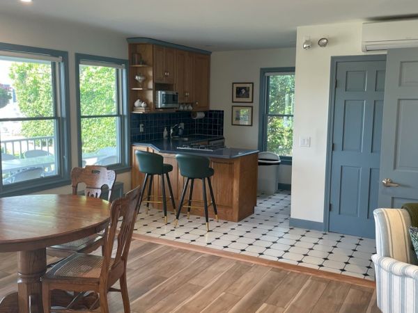 A cozy kitchen and dining area featuring wooden furniture, green bar stools, a small island, and large windows bringing in natural light.