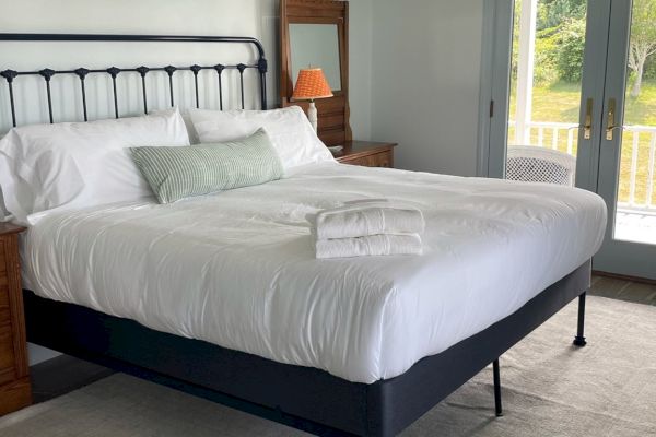 A neatly made bed with white linens, a green pillow, and folded towels on top, flanked by wooden nightstands, beside a glass door leading outside.