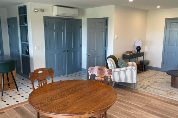 A cozy living space with wooden table and chairs, a striped armchair, small round table, and blue doors, adjacent to a kitchen area with barstools.