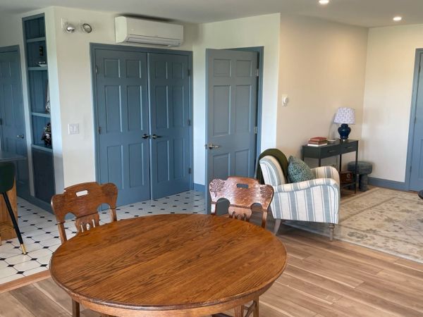 A cozy living space with wooden table and chairs, a striped armchair, small round table, and blue doors, adjacent to a kitchen area with barstools.
