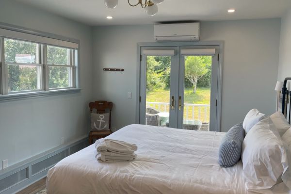 A well-lit bedroom with a large bed, folded towels, windows, glass doors leading outside, and a wall-mounted air conditioner.