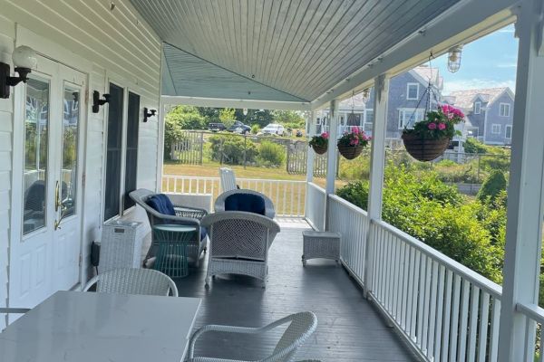 A cozy porch features wicker furniture, hanging flower baskets, and a white table with chairs, overlooking a tranquil outdoor view with nearby houses.