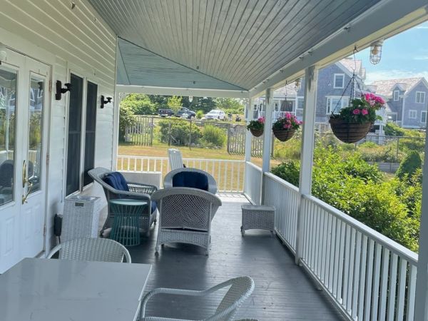A cozy porch features wicker furniture, hanging flower baskets, and a white table with chairs, overlooking a tranquil outdoor view with nearby houses.