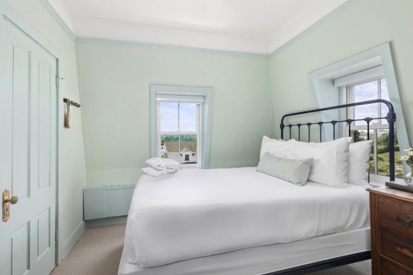 The image shows a cozy bedroom with pale green walls, a double bed, and two windows. There is a wooden nightstand with a lamp and decorative items.