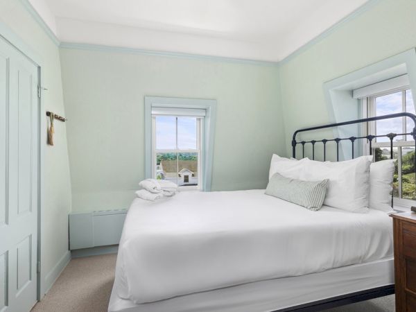 The image shows a cozy bedroom with pale green walls, a double bed, and two windows. There is a wooden nightstand with a lamp and decorative items.