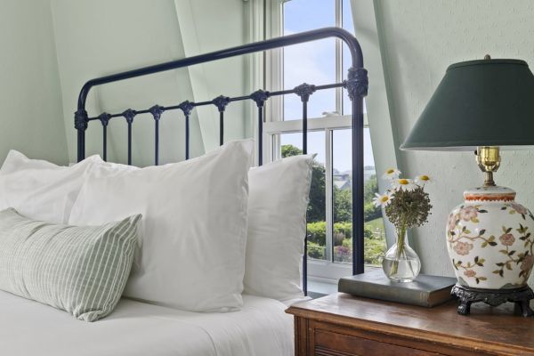 A neatly made bed with white pillows and a green pillow, next to a bedside table with a floral lamp, a vase of flowers, and a book by the window.