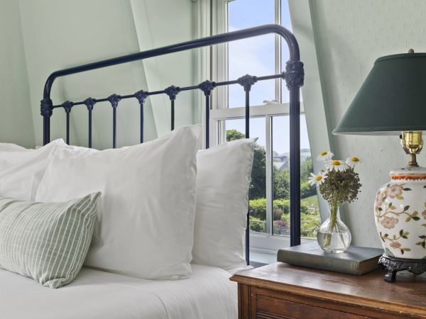 A neatly made bed with white pillows and a green pillow, next to a bedside table with a floral lamp, a vase of flowers, and a book by the window.