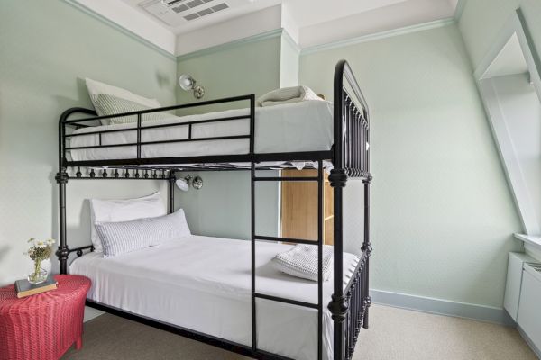 A neatly arranged bedroom with a black metal bunk bed, a red side table with a vase and a mirror on the wall, and a window with natural light.