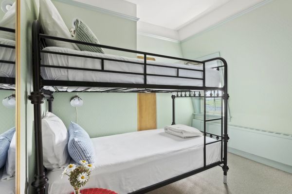 The image shows a room with a metal bunk bed, white sheets, pillows, a small bouquet of flowers, and light green walls.