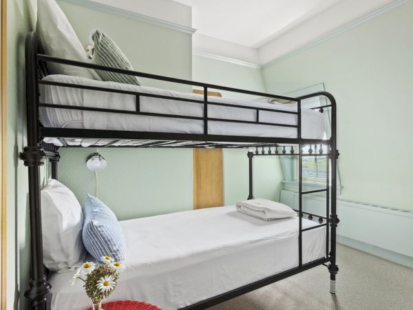 The image shows a room with a metal bunk bed, white sheets, pillows, a small bouquet of flowers, and light green walls.