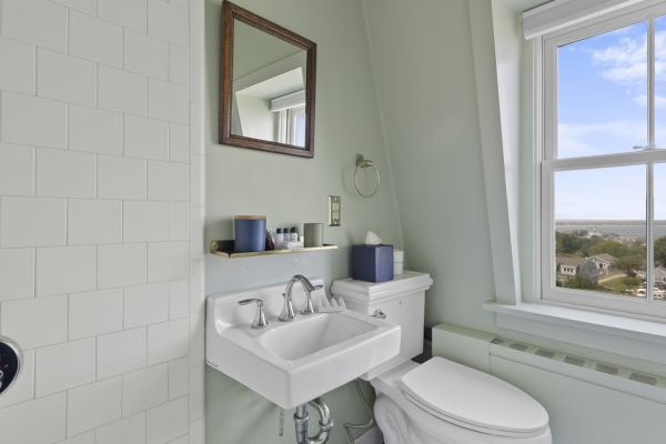 A bathroom with a sink, toilet, mirror, and window overlooking a landscape. There is a shelf above the sink with toiletries.