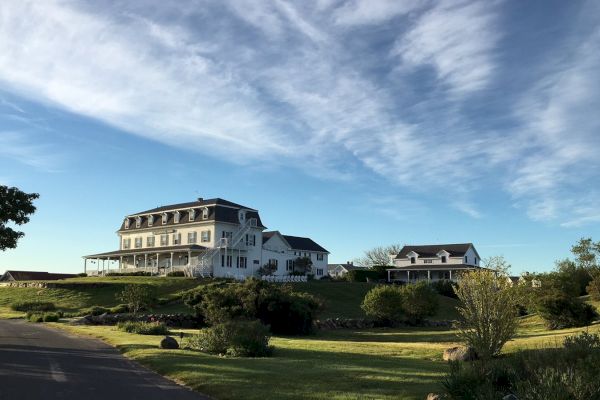 A large house with a wraparound porch sits on a hill, surrounded by greenery and a beautiful, partly cloudy sky.