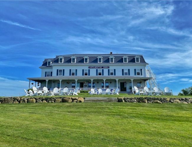 The image shows a large, multi-story white house with a wide porch, surrounded by green grass under a blue sky.